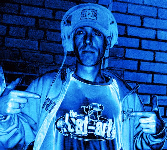DJ Natural Nate® in DC helmet and The-Lost-Art™ shirt pointing at camera under blue neon light against brick wall